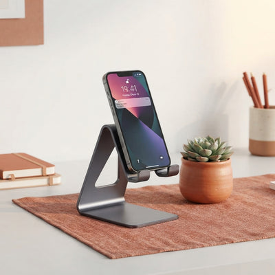 A dark gray aluminum phone stand on a desk with terracotta accents, holding a smartphone in a clean, professional workspace.